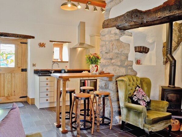 A rustic kitchen with a stone wall, wooden bar stools, a green armchair, and a vintage stove.