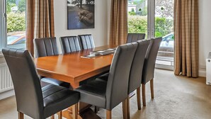 A dining room with a wooden table and six chairs, large windows with curtains, and a painting on the wall. - 4 bedroom accommodation in Keswick (Keswick)