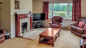 A living room with a fireplace, television, leather sofas, and a view of greenery outside. - 4 bedroom accommodation in Keswick (Keswick)