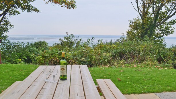 Una mesa de picnic de madera con una botella de vino y dos copas, frente a un área verde y vista al mar.