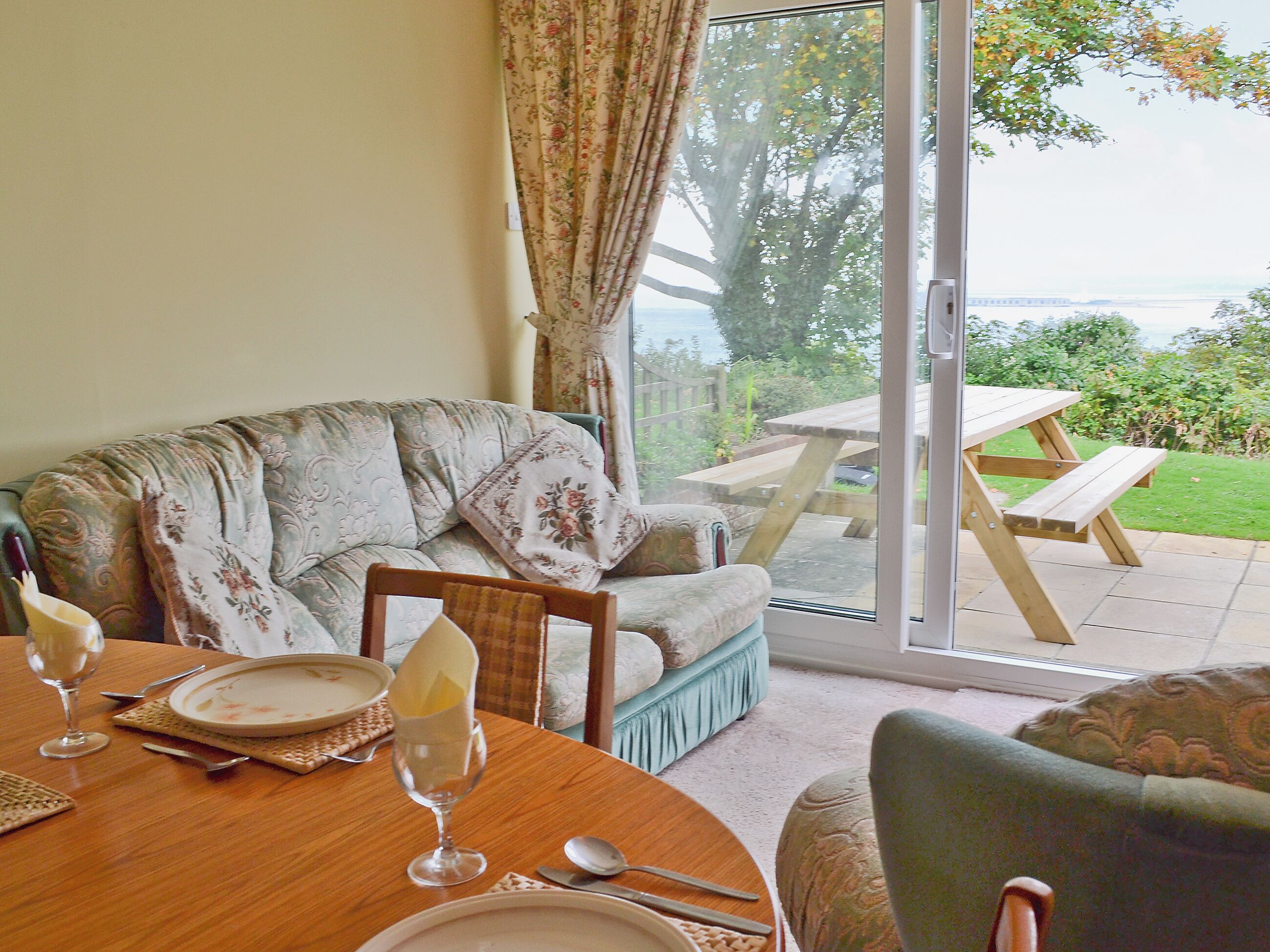 A dining area with a table set for two, a sofa, and a view of a garden and outdoor seating area.
