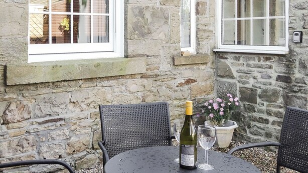 Ein Steinhaus mit zwei Fenstern in weißen Rahmen und einem Tisch mit Flasche und Glas.