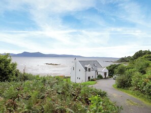 A coastal building with a paved driveway leading to a white entrance, surrounded by greenery and overlooking a body of water with distant landforms. - Macinnisfree Cottage (Saasaig, Teangue, Isle of Skye)