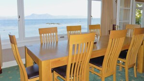 A dining area with a table and chairs, large windows with a view of the sea and mountains, and a carpeted floor. - Macinnisfree Cottage (Saasaig, Teangue, Isle of Skye)