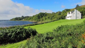 A coastal house with a grassy lawn, a view of the sea, and a hill with trees. - Macinnisfree Cottage (Saasaig, Teangue, Isle of Skye)