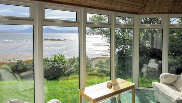 A room with a large window overlooking a coastal landscape, a wooden ceiling, and a small table. - Macinnisfree Cottage (Saasaig, Teangue, Isle of Skye)