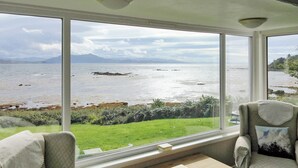 A room with a large window offering a view of the sea and distant mountains. There is a small table with a book on it. - Macinnisfree Cottage (Saasaig, Teangue, Isle of Skye)