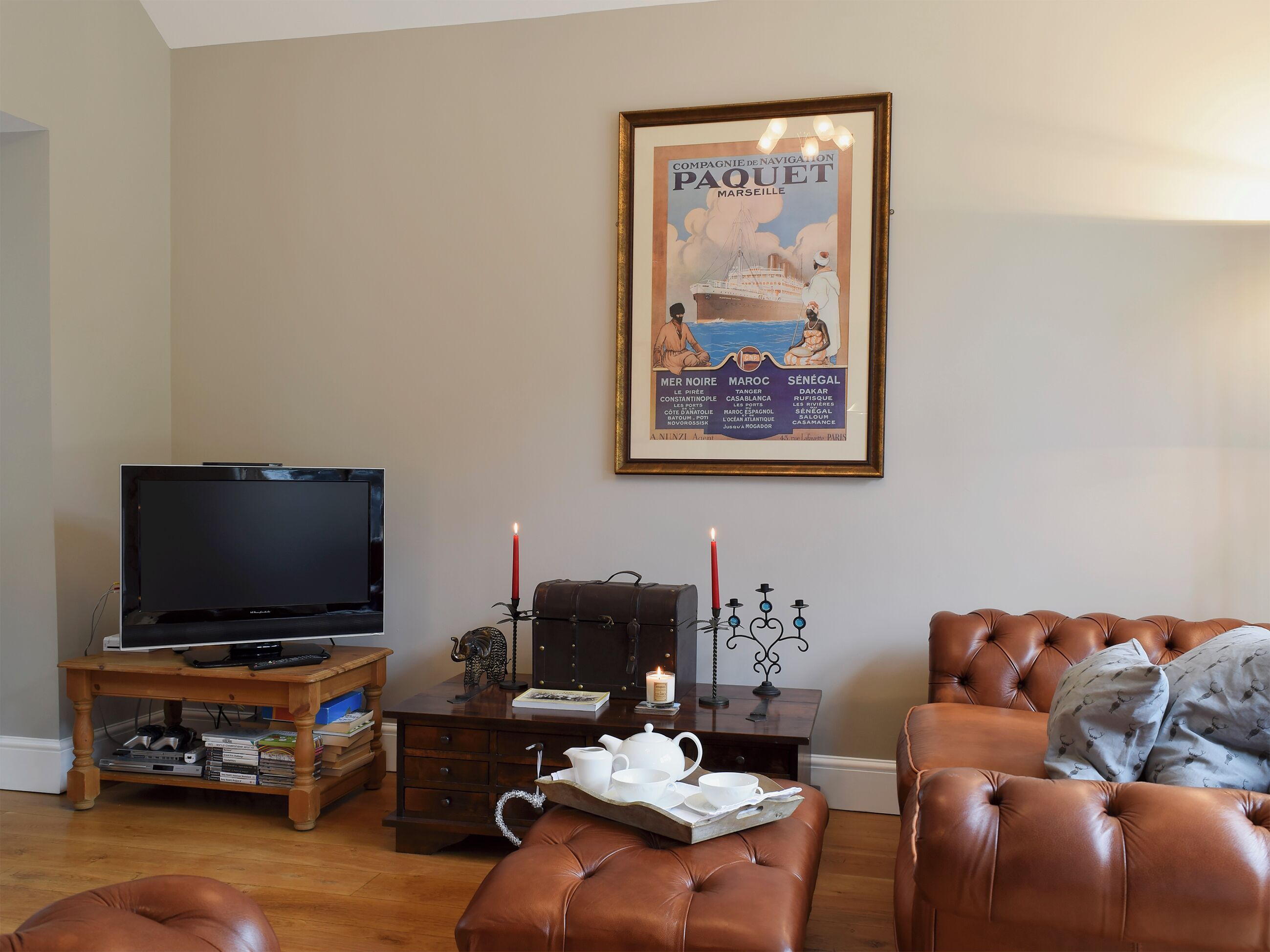 En stue med et fjernsyn, et indrammet plakat, et træ sofabord med stearinlys og en lædersofa.