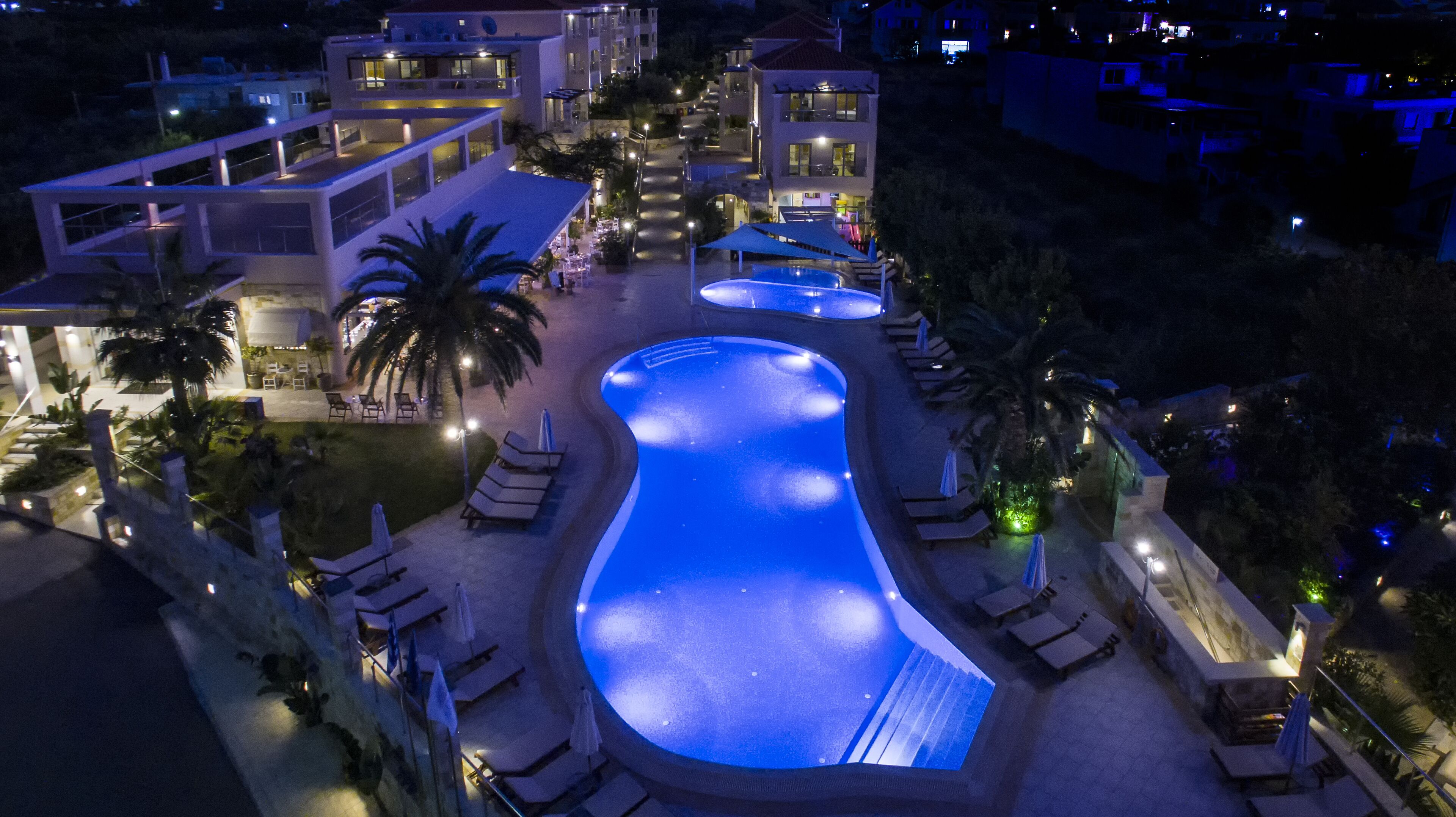 Une grande piscine illuminée, dotée de chaises longues et de parasols, entourée d’immeubles et de palmiers.