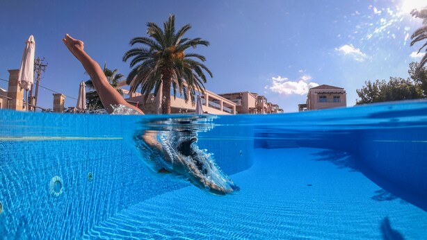 An underwater view of a swimming pool with clear blue water, palm trees, and buildings in the background. - Isida Hotel (Chania)
