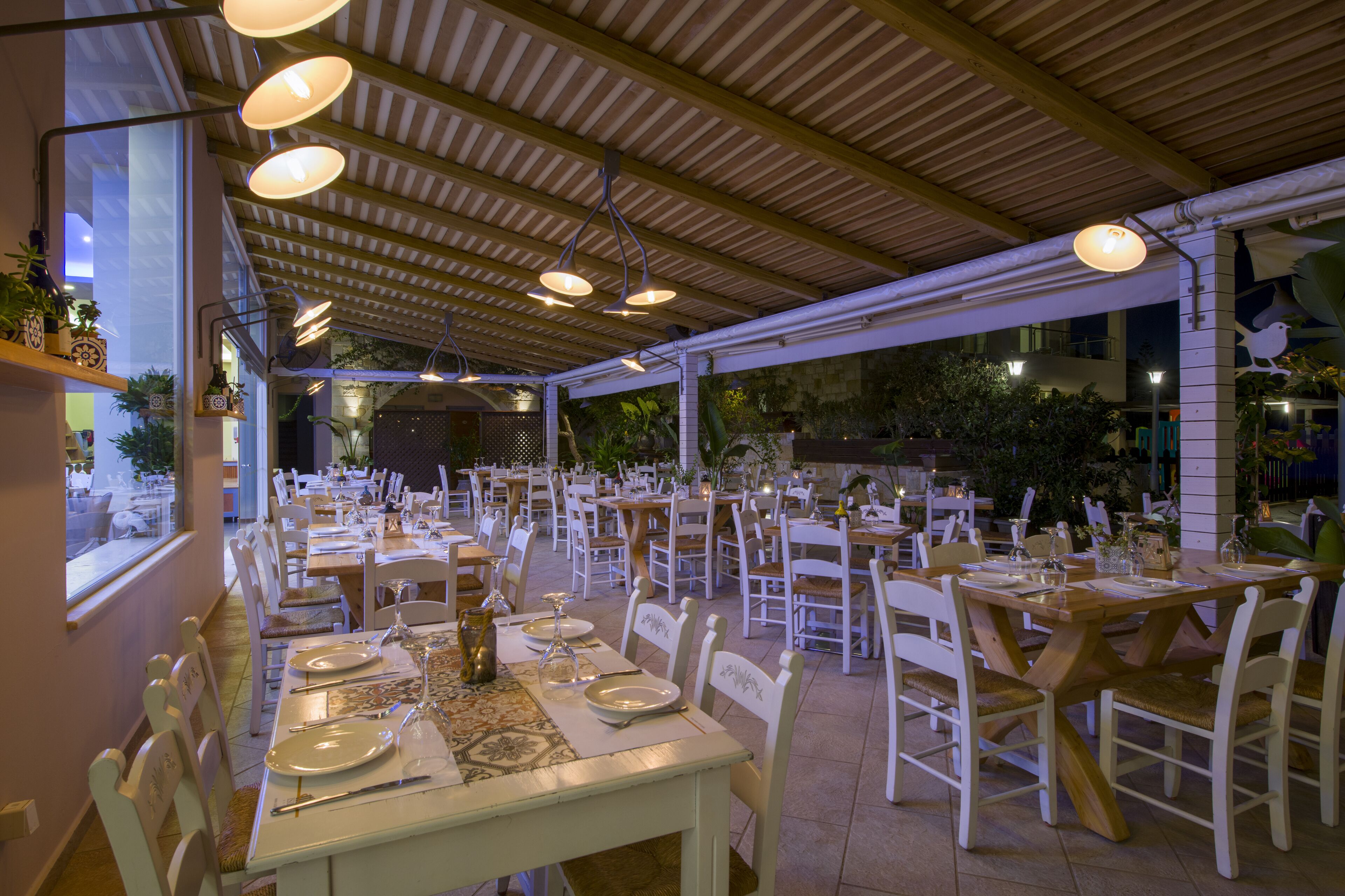 Un área de comedor al aire libre con mesas y sillas, cubierta por un techo de madera y rodeada de plantas y árboles.