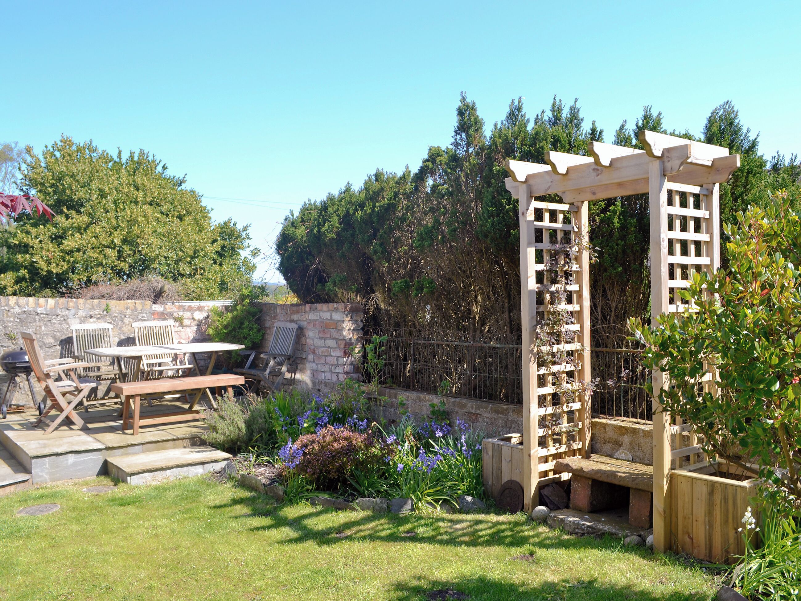 Eine Holz-Pergola mit einer rankenden Pflanze, eine Steinmauer und eine Terrasse mit Holzmöbeln.