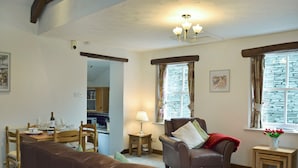Ein Wohnzimmer mit braunem Ledersofa, einem Holzesstisch mit Stühlen, einem kleinen Beistelltisch mit Lampe und einem Fenster mit Vorhängen.