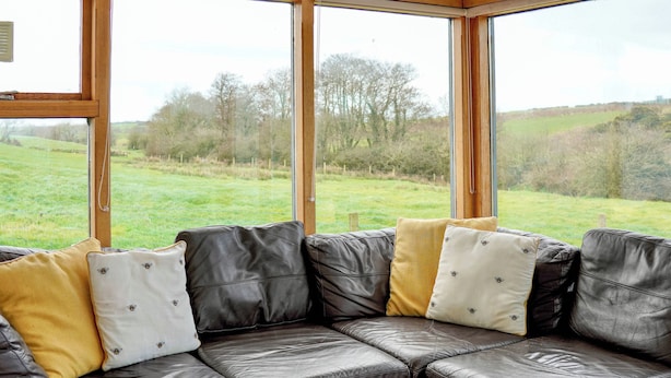 A sectional sofa with multiple cushions, a wooden table, and a view of a grassy field through large windows. - 2 bedroom accommodation in Tynlon, near Rhosneigr (Tynlon, near Rhosneigr)