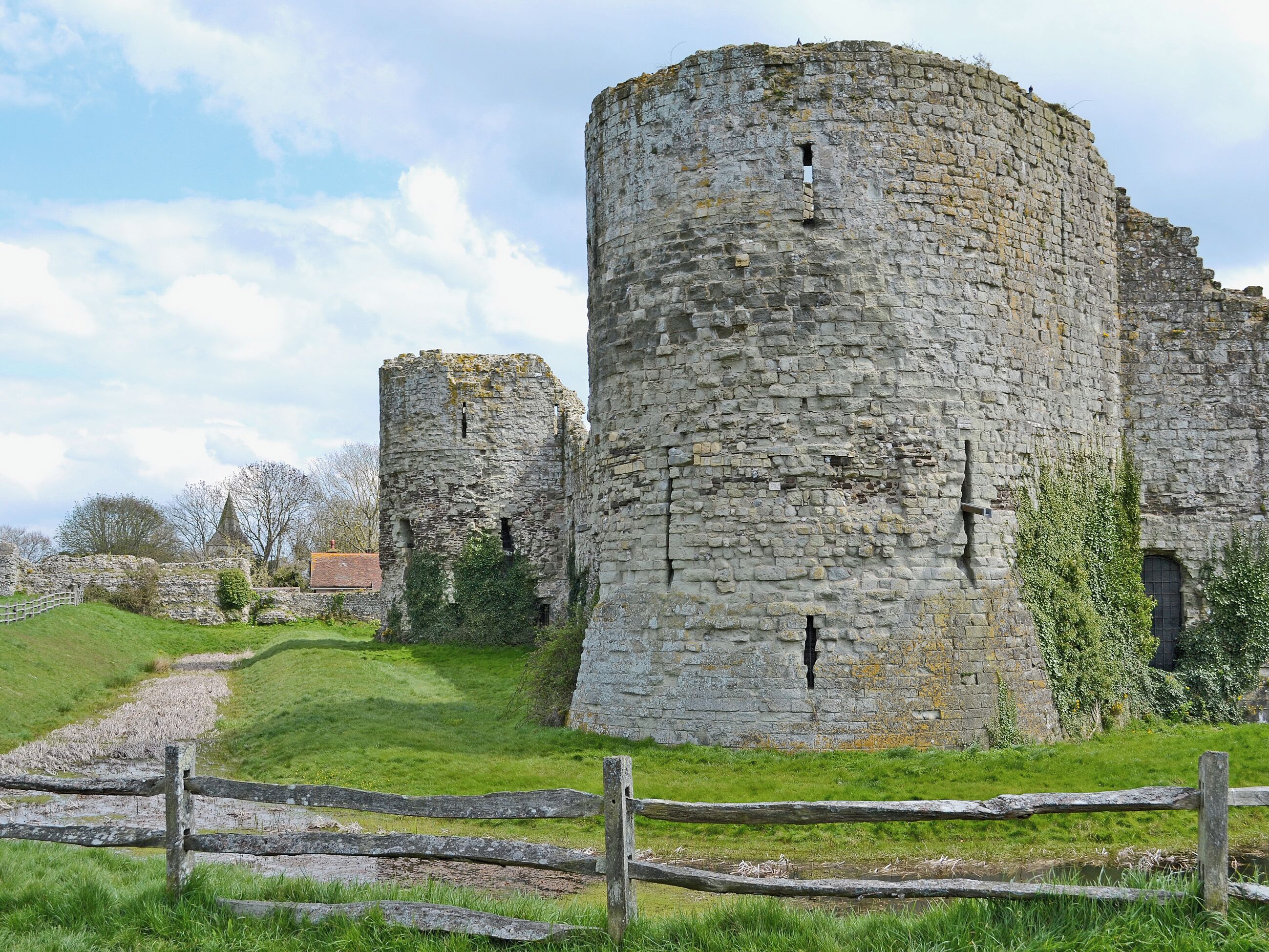Eine historische Burg aus Stein mit einem großen, zentralen Turm und kleineren Türmen, umgeben von einer grünen Wiese und einem Holzzaun.