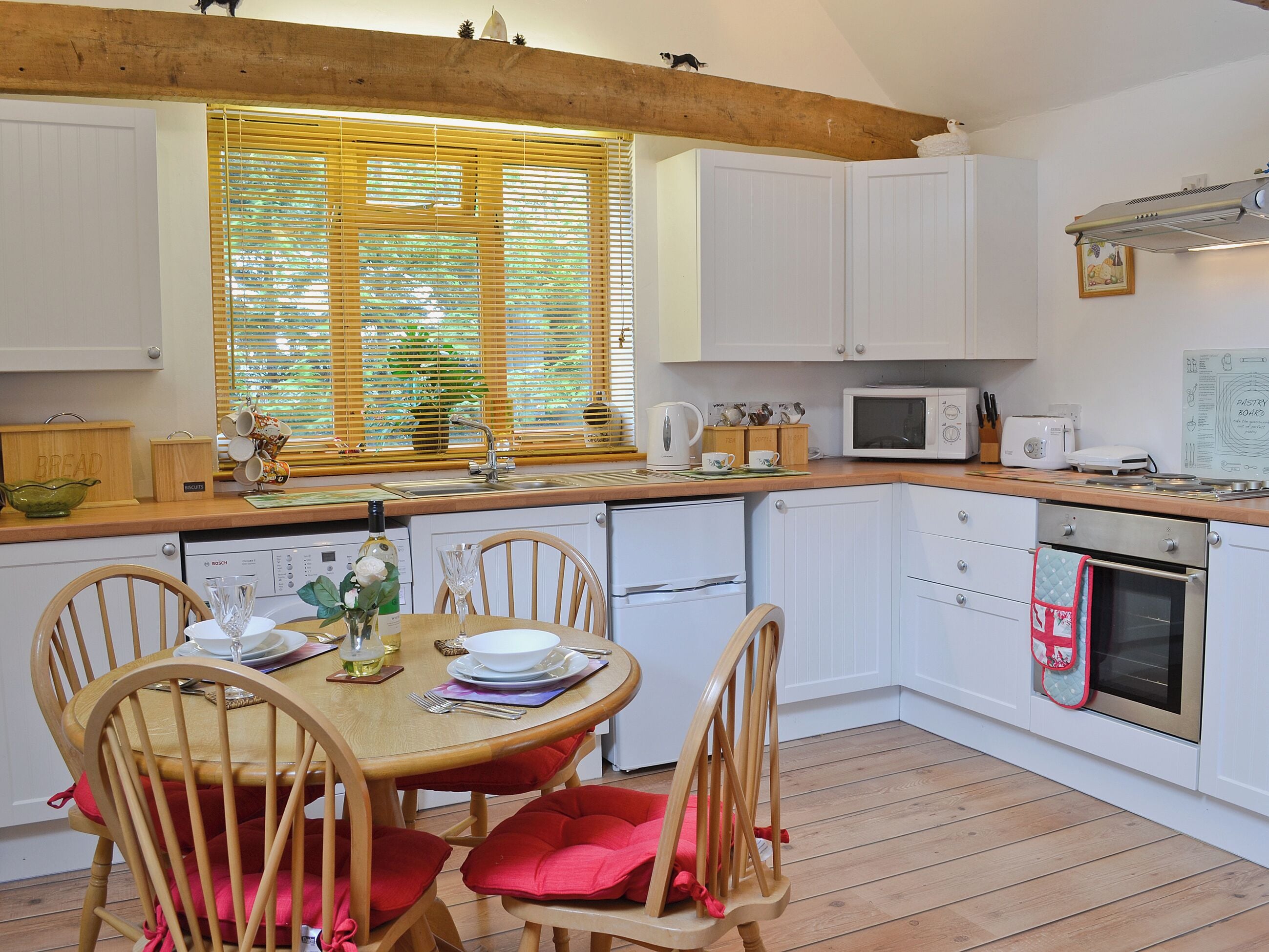 Une cuisine avec des meubles blancs, un plan de travail en bois, une table à manger avec des chaises et une fenêtre avec des stores.
