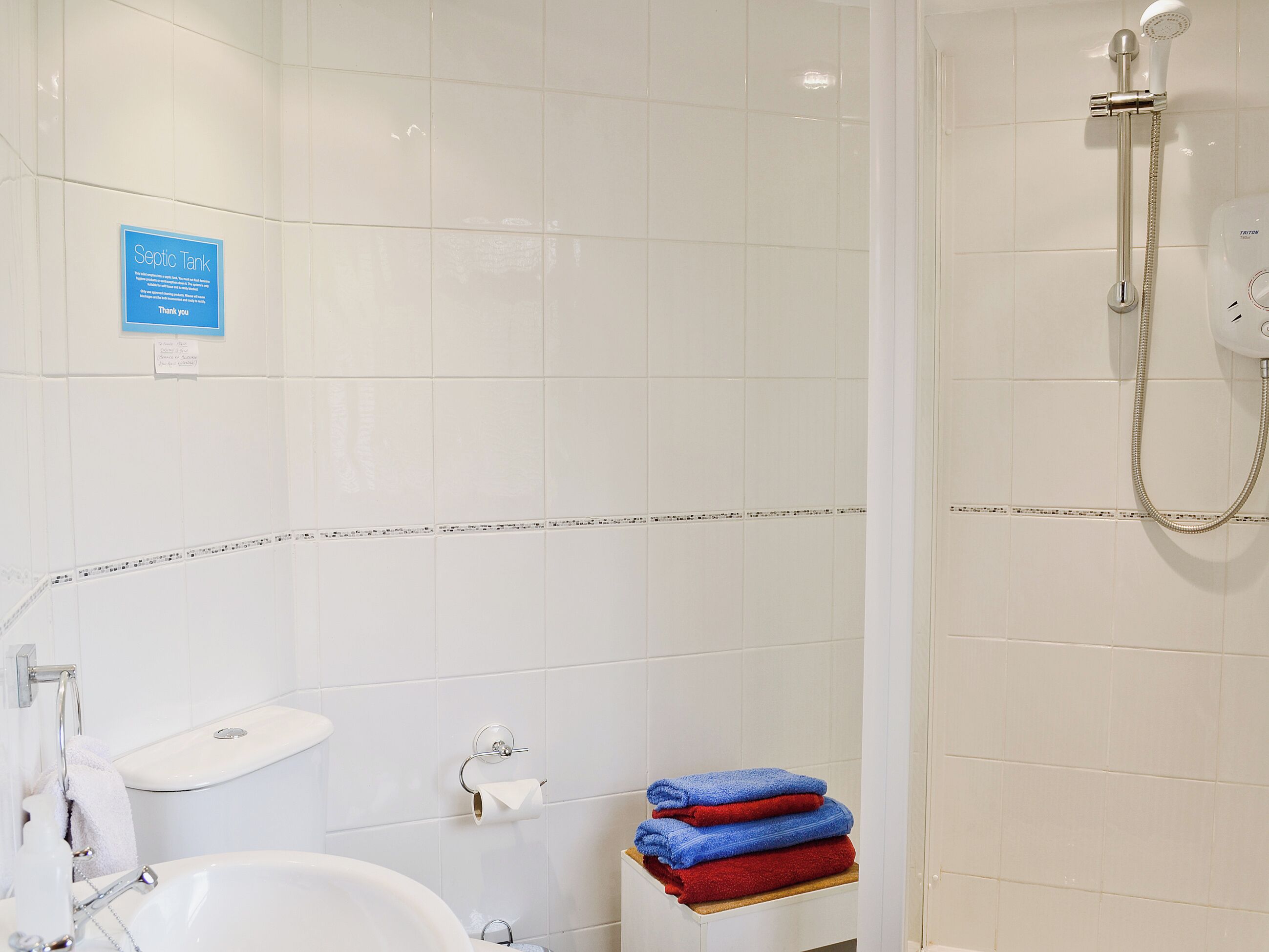 A bathroom with a white sink, toilet, and shower. There is a sign on the wall and a stack of towels.