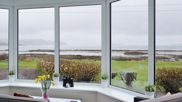 A conservatory with large windows overlooking a coastal landscape. Inside, there is a table with a vase of flowers and a pair of binoculars. - 2 bedroom accommodation in Lower Harrapool, near Broadford (Lower Harrapool, near Broadford)