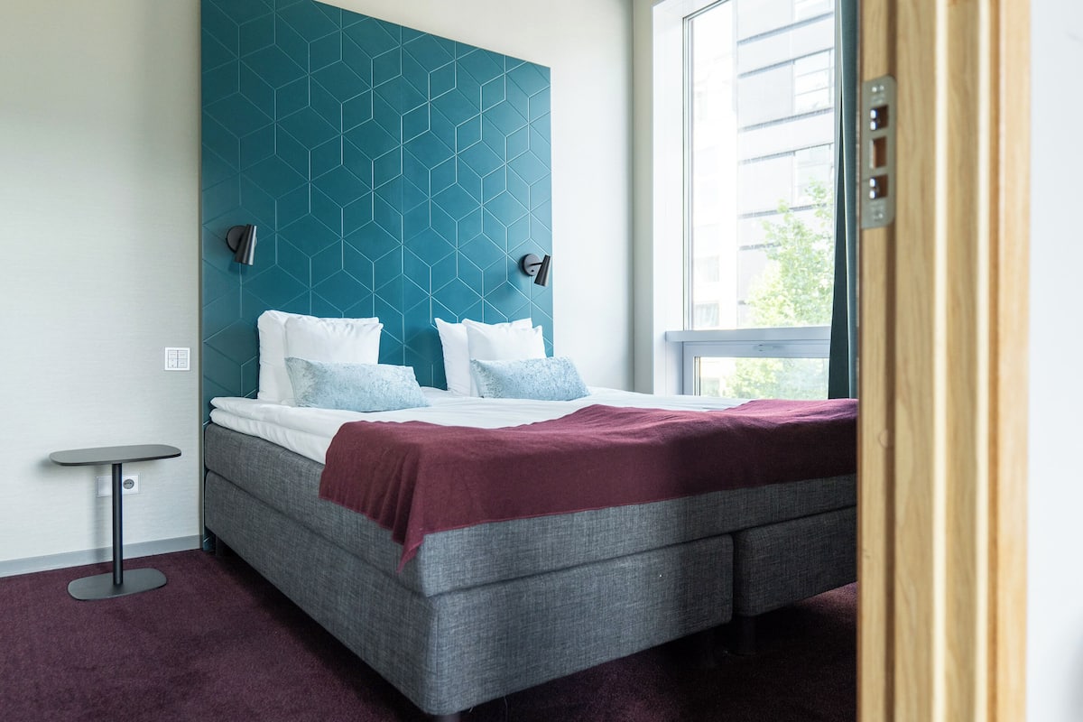 A modern bedroom with a geometric-patterned headboard, a grey bed with white and blue pillows, a red throw blanket, and a small bedside table.