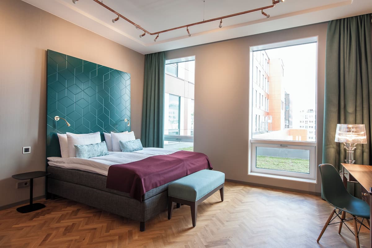 A modern hotel room with a large bed, a bench, a desk, and a view of the city through large windows.