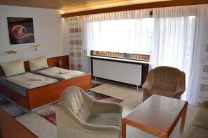 A hotel room with two beds, a sofa, a chair, a table, a radiator, and a window with curtains. - Hotel Garni Sebastian (Kirrweiler)