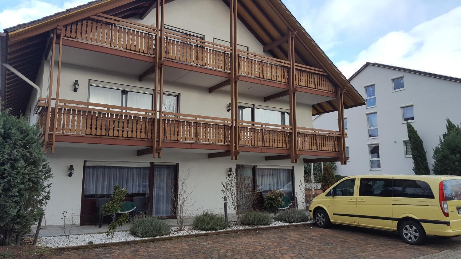 A multi-story building with balconies, a yellow car parked in front, and a garden area.
