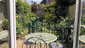 Une table ronde en métal, entourée de quatre chaises, se trouve dans un espace jardin agrémenté d’une clôture en bois et de verdure.