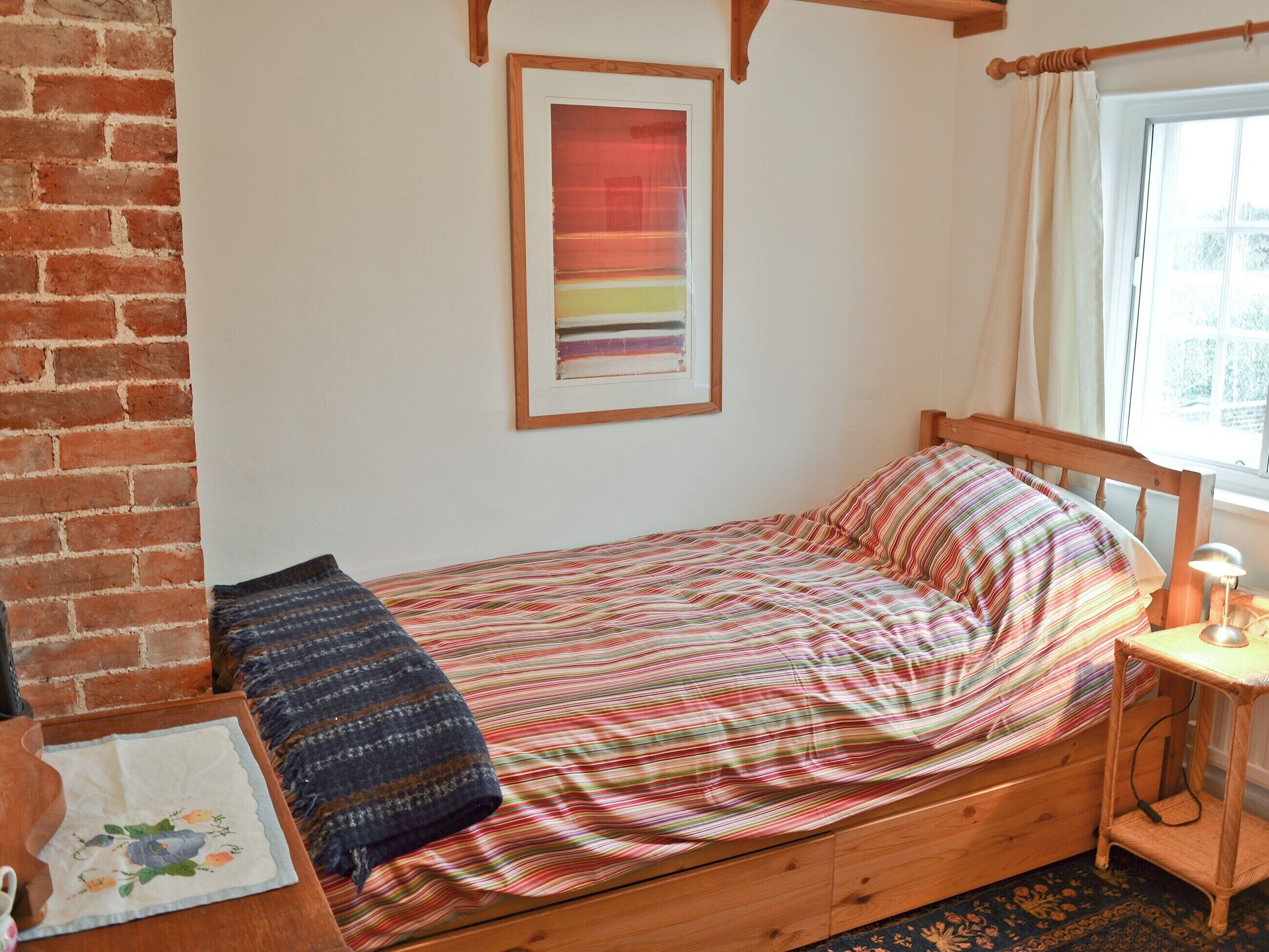 A single bed with a striped bedspread, a wooden headboard, a small bedside table with a lamp, and a framed abstract painting on the wall.