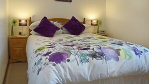 A neatly made bed with a floral bedspread, two purple pillows, a wooden headboard, a nightstand with a lamp, and a wall-mounted picture. - Elm Court 18 has the advantage of being a semi-detached house. (Keswick)