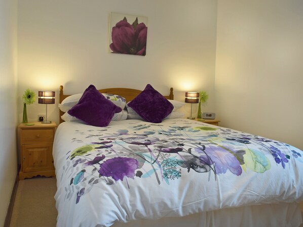 A neatly made bed with a floral bedspread, two purple pillows, a wooden headboard, a nightstand with a lamp, and a wall-mounted picture. - Elm Court 18 has the advantage of being a semi-detached house. (Keswick)