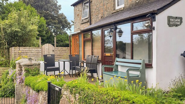 A stone cottage with a patio, outdoor seating, and a sign reading "Trecarne Cottage".