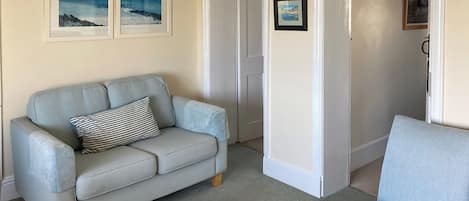 A living room with a light blue sofa, a striped pillow, and a light blue armchair. There are two framed pictures on the wall, a door, and a carpeted floor.