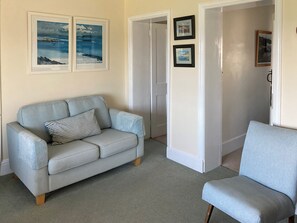 A living room with a light blue sofa, a striped pillow, and a light blue armchair. There are two framed pictures on the wall, a door, and a carpeted floor.
