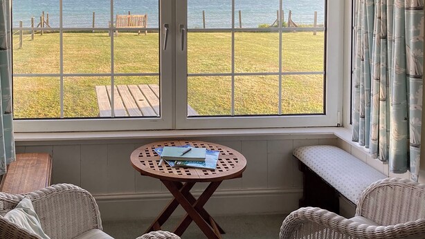 A room with a large window overlooking a grassy area and the sea, a small round table with books, and wicker furniture. - 3 bedroom accommodation in Downderry, near Looe (Downderry, near Looe)