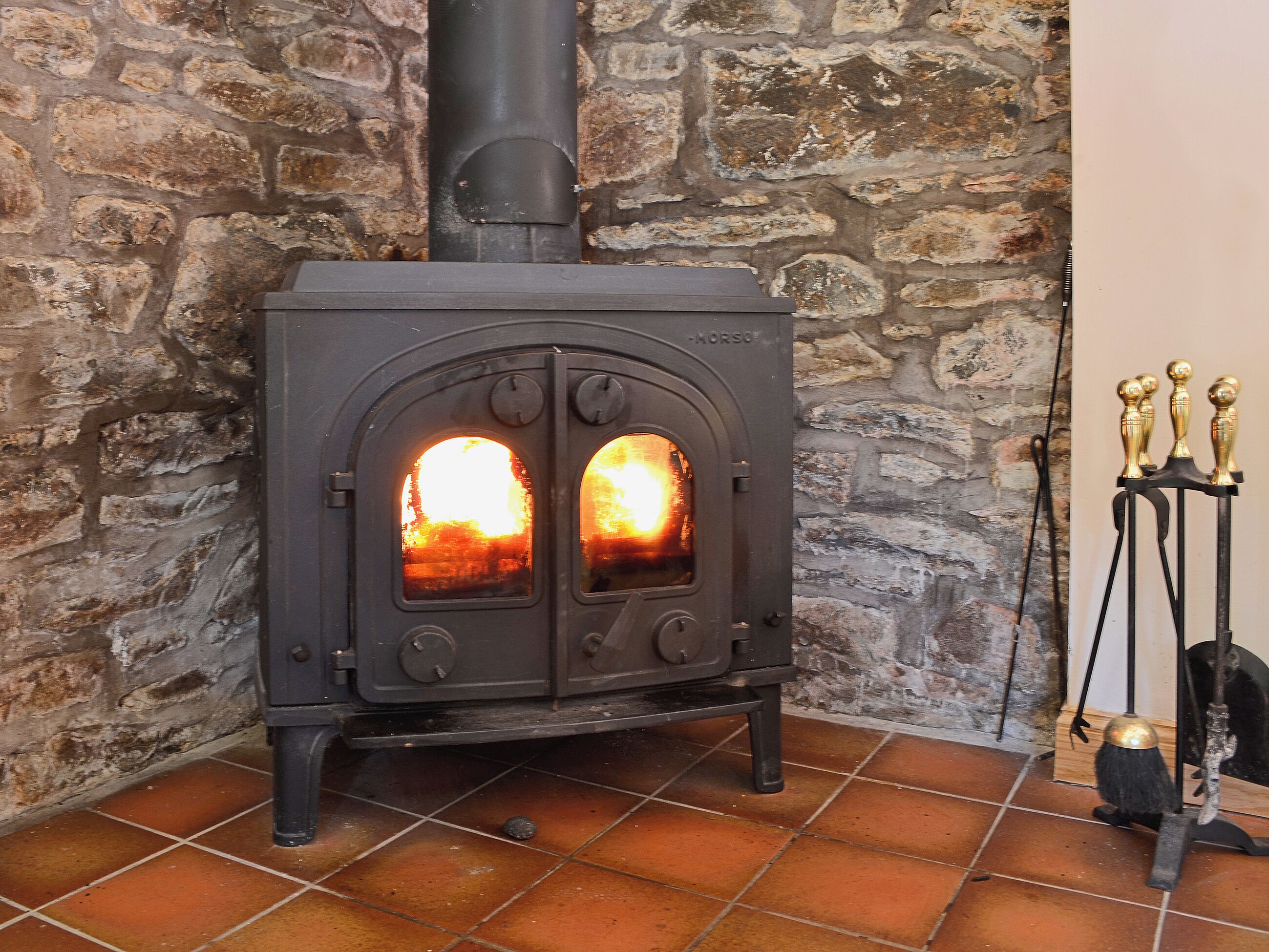 A wood-burning stove with two visible fireboxes, set against a stone wall background.