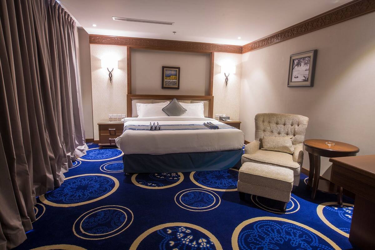 Habitación de hotel con una cama grande, una silla, una mesita pequeña y una alfombra azul estampada.
