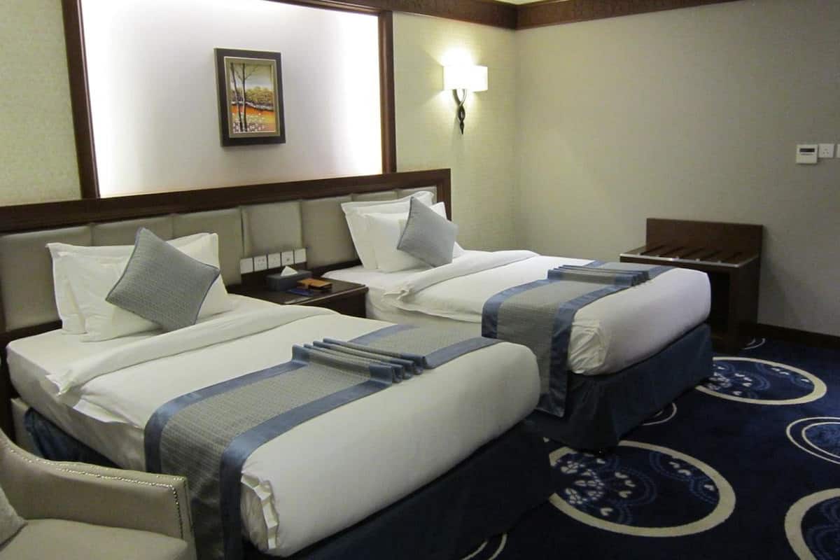 Una habitación de hotel con dos camas, una mesita de noche, una silla, una lámpara de pared y un cuadro enmarcado.