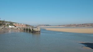 Un paysage côtier avec une jetée s’avançant dans des eaux calmes, une plage de sable et des bâtiments situés sur une colline.