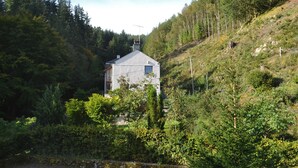 A small, single-story house surrounded by dense forest, with a sloped roof and a chimney.