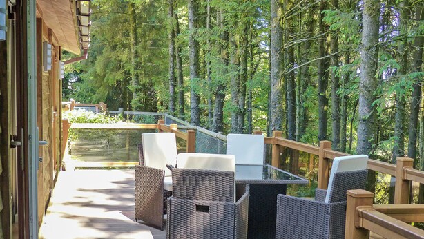 An outdoor deck with wicker furniture and a glass table, surrounded by a wooden railing and dense forest. - 2 bedroom accommodation in Clatworthy, near Williton (Clatworthy, near Williton)