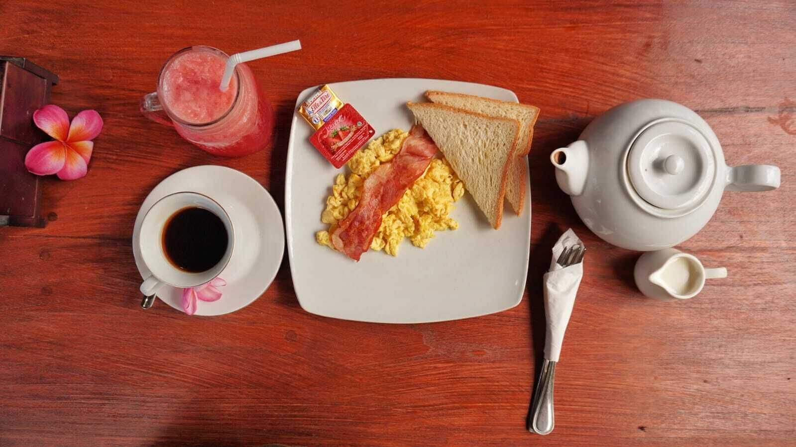 A breakfast setting with scrambled eggs, bacon, toast, a cup of coffee, a teapot, and a small container of jam on a wooden table.