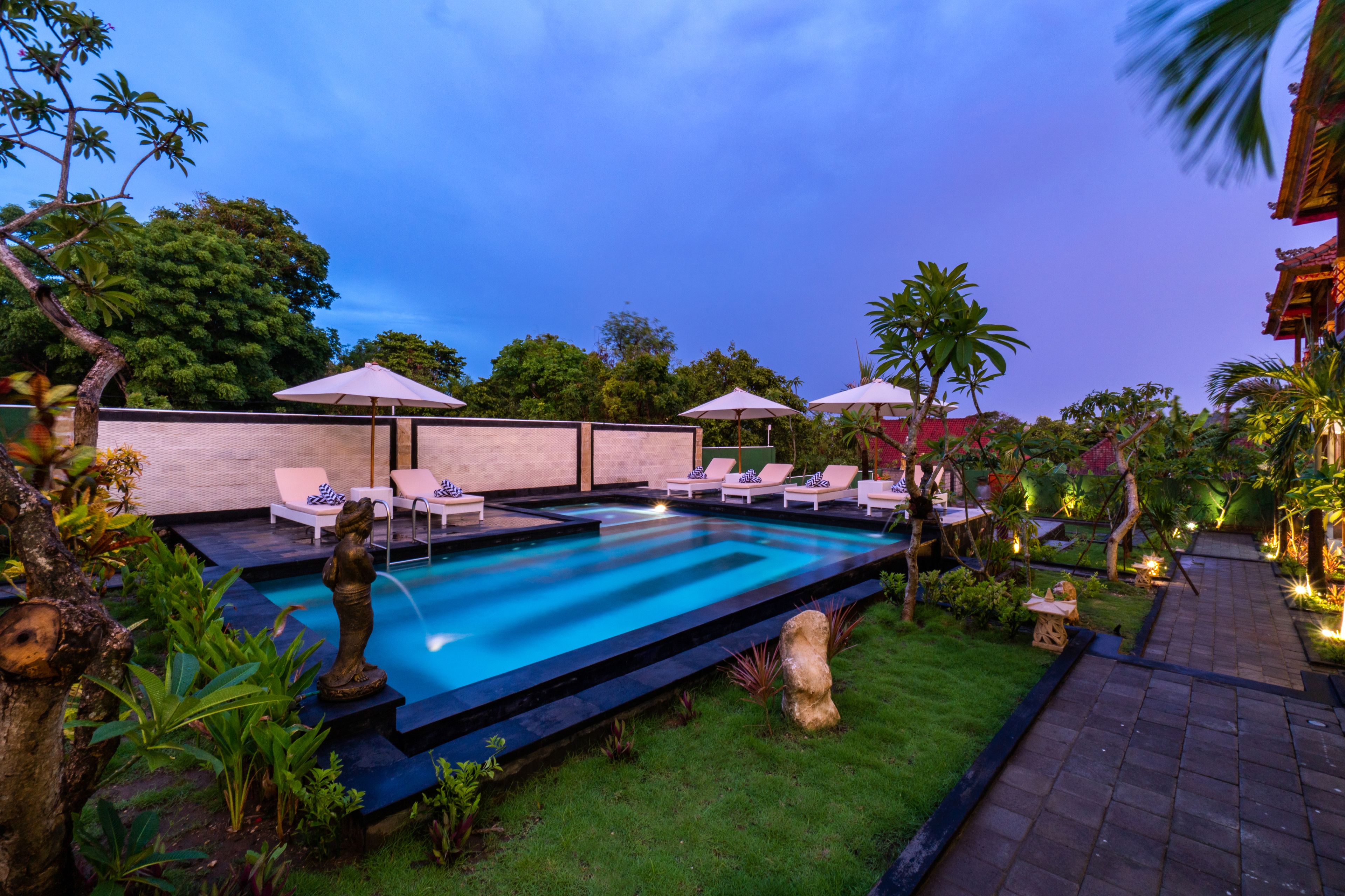 A swimming pool with lounge chairs and umbrellas, surrounded by tropical plants and a brick pathway.