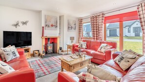 A cozy living room with a fireplace, red leather sofas, a flat-screen TV, and a large window with curtains.