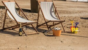 Zwei Strandstühle mit gestreiften Kissen, eine Weinflasche, ein Glas und Strandspielzeug am sandigen Strand.