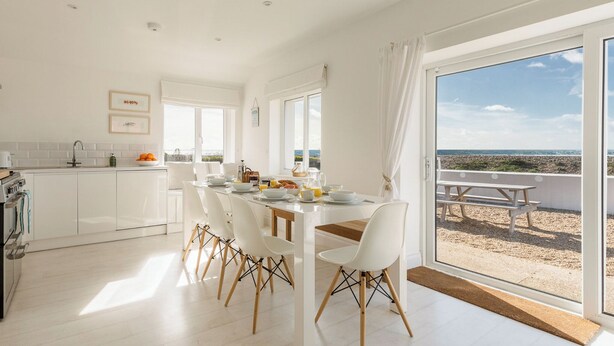 Eine moderne Küche mit weißen Schränken, ein Essbereich mit weißen Stühlen und einem Holztisch sowie eine große Schiebeglastür mit Blick auf den Strand.