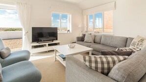 Ein modernes Wohnzimmer mit Flachbildfernseher, einer Couch, einem Couchtisch und einem Fenster mit Blick auf einen Strand.