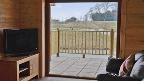 Ein Holzhäuschen-Innenraum mit einem Fernseher auf einem Holzständer, einer Couch mit Kissen und einem Balkon mit Holzgeländer.