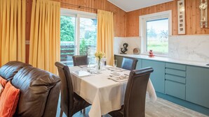 Un coin repas avec une table dressée pour quatre personnes, des murs en bois, une fenêtre avec des rideaux et un espace cuisine doté de meubles de couleur turquoise.