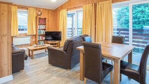 Un salon confortable avec un canapé, un téléviseur, une table basse et une table à manger avec des chaises. On y trouve une bibliothèque remplie de livres et une fenêtre avec des rideaux.