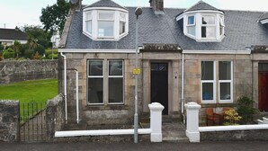 A stone building with two bay windows, a black door, and a small porch with wooden chairs. - 1 bedroom accommodation in Millport, Isle of Cumbrae (Millport, Isle of Cumbrae)